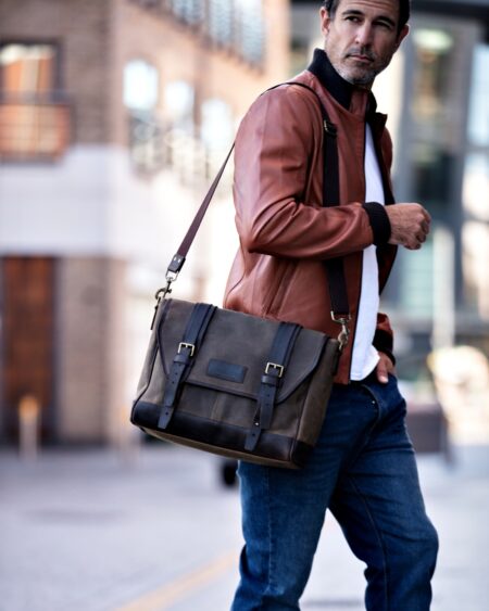 A middle-aged man in a brown leather jacket walking down a city street, carrying a vintage dark olive Builford Bohemian waxed canvas messenger bag.
