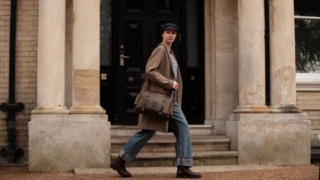 A three-quarter back view of a person with dark hair in a ponytail, wearing a beige trench coat, walking towards a grand, historical stone building. They are carrying a dark oak Builford Classic Hobo Messenger Bag made of texturized British original waxed canvas with brown leather straps, slung over their left shoulder. The setting is a paved courtyard on a partly cloudy day.